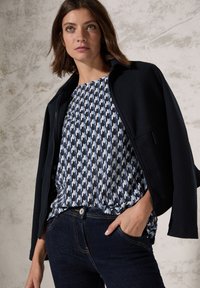Veste zippée navy sur un haut à motif bleu avec un design pied-de-poule, assortie à un jean en denim sombre. Textures lisses et forme ajustée.