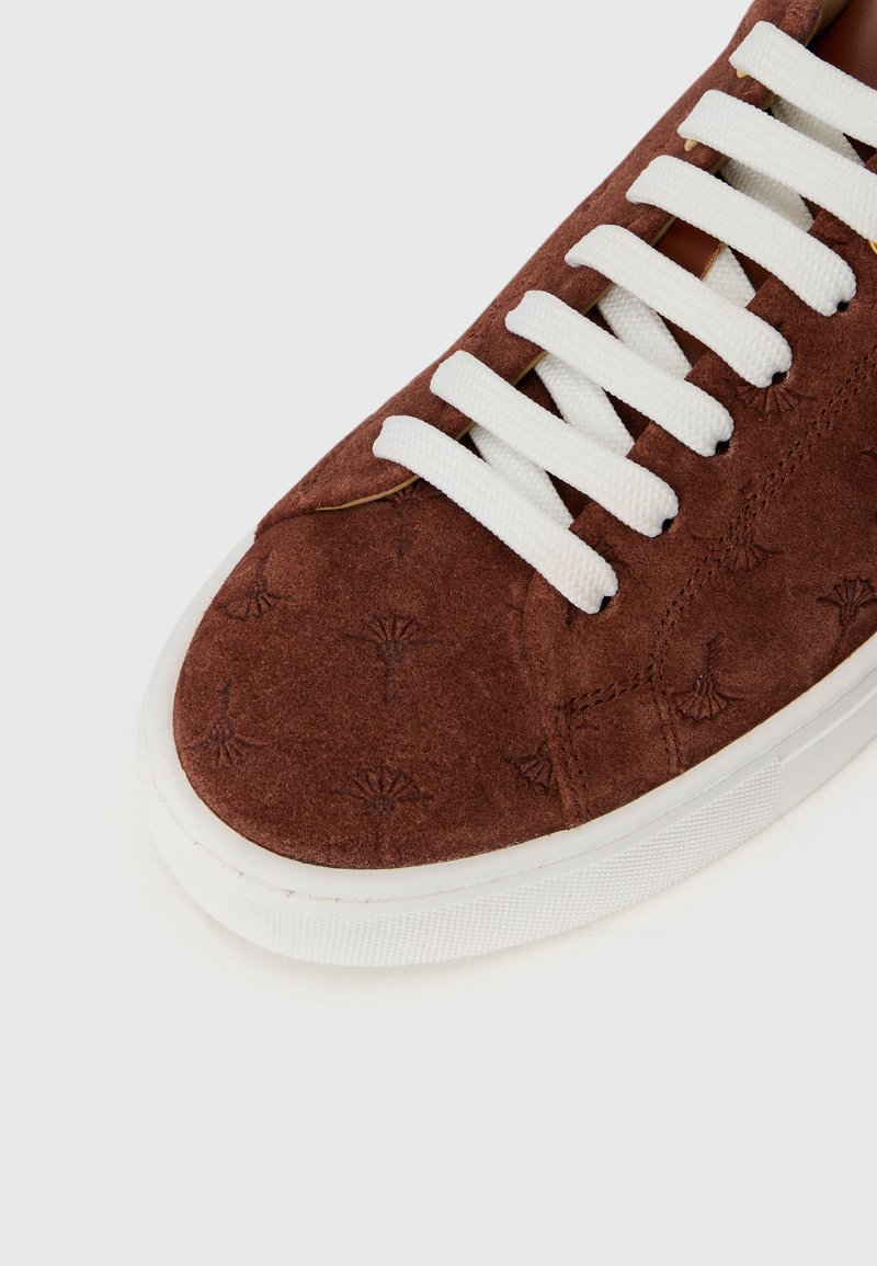 Braune Wildledersneaker mit weißen Schnürsenkeln, dezentem embossed Muster und weißer Gummisohle auf einem weißen Hintergrund.