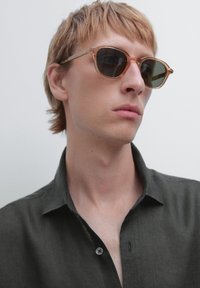 Heldere, beige zonnebril met een klassieke vorm ligt op een neutraal gekleurde shirt, accentueert door een enkele knop en een zachte textuur.