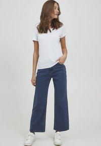 Balta kokvilnas T-krekla ar īsām piedurknēm kombinācija ar augsta jostasvieta platām zilām džinsu biksēm. Modelis valkā baltas sporta apavus.