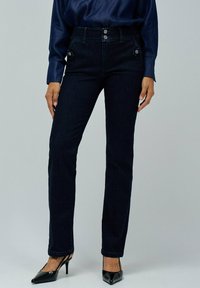 Vaqueros de mezclilla azul oscuro con cintura alta, cuatro bolsillos y detalles de botones metálicos. Combina con zapatos de tacón negros y una blusa azul marino.