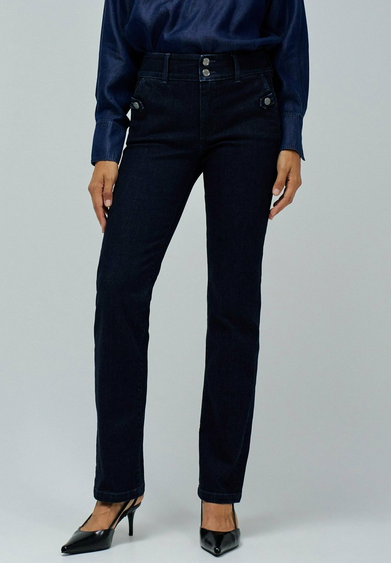 Vaqueros de mezclilla azul oscuro con cintura alta, cuatro bolsillos y detalles de botones metálicos. Combina con zapatos de tacón negros y una blusa azul marino.