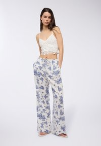 Top corto in pizzo bianco con spalline sottili, abbinato a pantaloni a gamba larga con motivo floreale blu su uno sfondo chiaro.