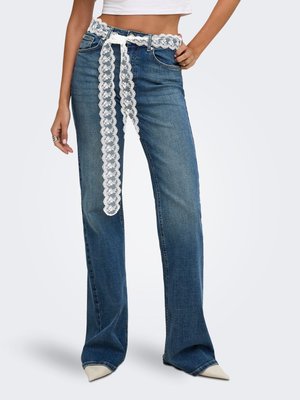 Femme portant un jean bleu droit avec ceinture en dentelle blanche et chaussures blanches pointues, crop top visible à la taille.