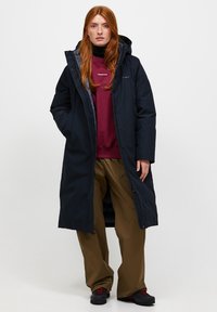 Lange marineblauwe parka met een capuchon, met een ritssluiting en zijzakken. G gedragen over een kastanjebruine sweatshirt en een paarse broek. Zwarte schoenen.