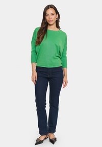 Grön stickad tröja med en avslappnad passform och trekvartsärmar, kombinerad med mörka denimjeans och svarta spetsiga flats.