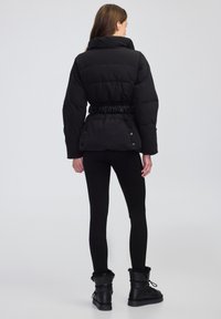 Donna che indossa un giubbotto imbottito nero con vita arricciata, leggings neri e stivaletti neri foderati in pelliccia, in piedi di spalle su uno sfondo neutro.