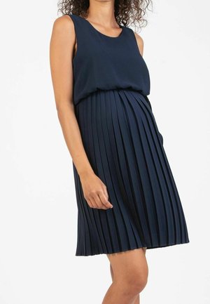Femme portant une robe bleu marine sans manches avec jupe plissée, debout sur fond blanc, recadrée aux épaules et aux genoux.