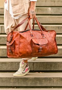 Duża brązowa skórzana torba podróżna z dwiema przednimi kieszeniami, srebrnymi okuciami i wyraźną fakturą, noszona przez osobę na kamiennych schodach.