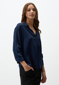 Blusa a maniche lunghe blu navy con colletto, caratterizzata da un ricamo sottile. Realizzata in un materiale leggero e liscio. Vestibilità comoda.