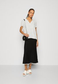 Top bianco a maniche corte con motivo a macchie nere; gonna nera alla caviglia con orlo ondulato; mules bianche con platform; piccola borsa nera.