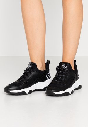 Gambe con indosso sneaker chunky nere e bianche con lacci neri e audace logo bianco sui lati e sulla linguetta su sfondo bianco.