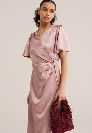 Sieviete, tērpusies zīdaini rozā apsaiņošanu kleitā ar plandošām piedurknēm, turot teksturētu bordo ziedu somu uz vienveidīga bēša fona.