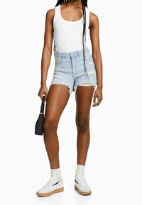 Ljusblå denimshorts med fransade kanter och slitna detaljer, parat med en vit tanktop och vita sneakers, hållande en liten svart väska.