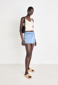 Top de tip tank beige deschis cu dantelă la margini, asortat cu o fustă mini din denim albastru. Îmbrăcat cu sandale bej fără toc. Modelul stă în fața unui fundal simplu.