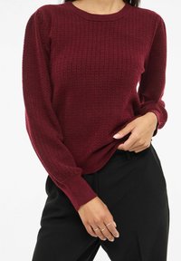 Burgundowy sweter dziany z teksturowanym wzorem, ściągaczami na rękawach i okrągłym dekoltem, w połączeniu z czarnymi spodniami. Ręce spoczywają na pasku spodni.