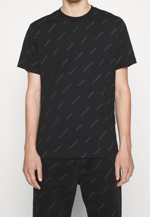 Personne portant un t-shirt noir uni à manches courtes et un pantalon noir, debout sur un fond blanc.