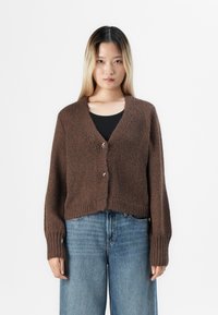 Kvinne som står mot hvit bakgrunn iført en brun strikket cardigan med sølvspenner over svart topp og løse blå jeans.