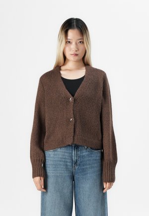 Kvinne som står mot hvit bakgrunn iført en brun strikket cardigan med sølvspenner over svart topp og løse blå jeans.