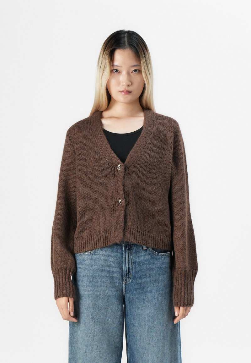 Kvinne som står mot hvit bakgrunn iført en brun strikket cardigan med sølvspenner over svart topp og løse blå jeans.