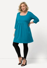 Tyrkysové šaty s čtvercovým výstřihem a tříčtvrtečními rukávy, s nařaseným pasem, v kombinaci s černými legíny a černými sandály na podpatku. Hladká textura.