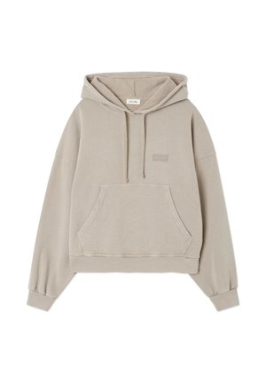 Personne portant un sweat à capuche ample gris clair et un pantalon large court associé à des bottines couleur sable, debout devant un fond uni.