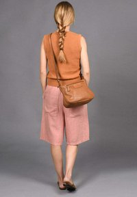 Tan mouwloze trui, roze linnen culottes en een bruine geperforeerde schoudertas. Model staat blootsvoets tegen een grijze achtergrond.