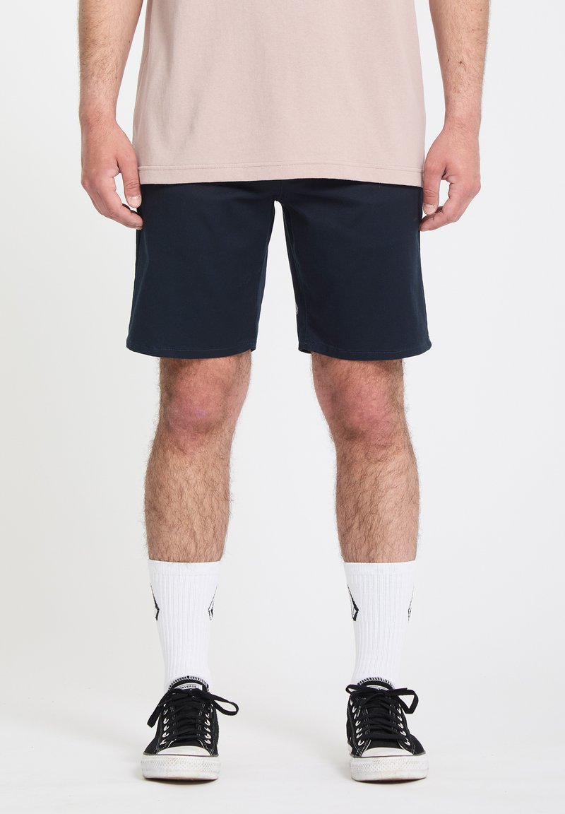 Άνδρας που φοράει μπεζ πουκάμισο, σορτς ναυτικού μπλε, λευκές κάλτσες και μαύρα αθλητικά παπούτσια, στέκεται μπροστά σε απλό λευκό φόντο.