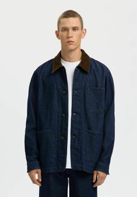 Jeune homme portant une veste en jean foncé avec un col marron et de grandes poches avant, debout devant un fond blanc uni.