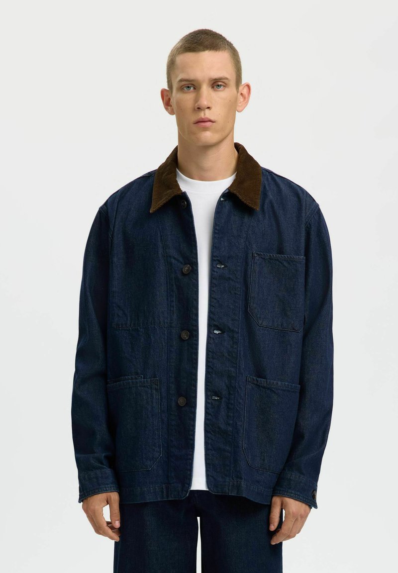 Jeune homme portant une veste en jean foncé avec un col marron et de grandes poches avant, debout devant un fond blanc uni.