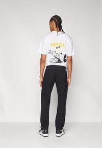 La camiseta blanca presenta un diseño con un personaje y texto en amarillo. Los pantalones cargo negros con bolsillos laterales y las zapatillas negras completan el atuendo.