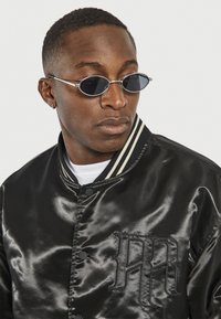 Chaqueta bomber de satén negro con detalle de logo, presenta un cuello a rayas y botones. Unas gafas de sol redondas y tintadas completan el look.