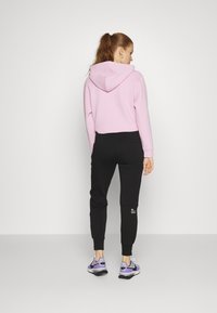 Rosa hettegensere med baklomme, kombinert med svarte joggebukser med en liten logo. Modellen har på seg lilla og svarte sneakers.