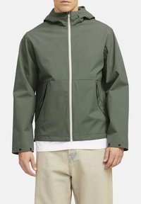 Giacca impermeabile verde con cappuccio, cerniera bianca, due tasche frontali, polsini elasticizzati e taglio dritto. Indossata sopra una camicia bianca.