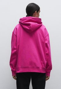 Růžová oversized mikina s kapucí na stažení, vroubkovanými manžetami a lemem, vyrobená z měkké tkaniny s hladkou strukturou. Zadní pohled.