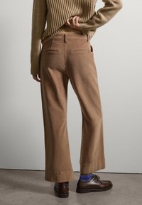 Los pantalones de pana marrón de pierna ancha presentan bolsillos traseros y una cintura de altura media, combinados con zapatos oscuros y calcetines multicolores.