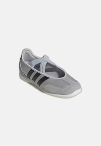 Sneaker stile Mary Jane grigio chiaro con strisce nere e cinturini elastici incrociati su sfondo bianco.