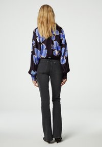Bloemige blouse in donkerpaars met grote blauwe en crèmekleurige bloemen, gecombineerd met zwarte flared jeans. Gevonden vanuit de achterkant, waarbij de haardracht en de pasvorm van de outfit zichtbaar zijn.