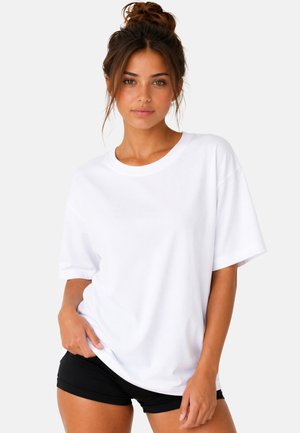 Jeune femme aux cheveux bruns attachés en chignon, portant un t-shirt blanc ample et un short noir, debout devant un fond clair uni.