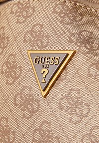 Tissu marron texturé avec un motif en relief subtil. Présente une plaque logo en or en forme de triangle avec du texte brun foncé "GUESS USA" et un point d'interrogation.