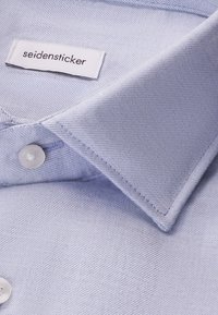Camicia azzurra chiara con texture liscia, colletto a punta e sottile trama diagonale. Presenta un'etichetta con la scritta "seidensticker."