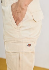 Pantalons cargo beiges en tissu texturé. Comprend une poche latérale et un écusson logo. Le design inclut une fermeture à bouton et des poches à plat.