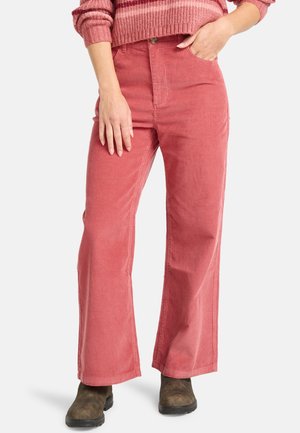 Nainen, jolla on ylipolviseen vyötäröön ulottuvat leveälahkeiset vaaleanpunaiset corduroy-housut, ruskeat nilkkurit ja vaaleanpunainen raitahelmalla varustettu neulepaita, ja toinen käsi taskussa.