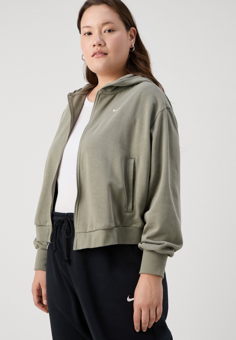 Sudadera con cremallera color verde oliva hecha de material suave, con capucha, bolsillos laterales y un pequeño logotipo blanco de Nike en el pecho. Combínala con pantalones negros.