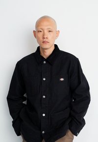 Black work jacket made of cotton with a buttoned front, two chest pockets, and a corduroy collar. Features silver hardware and logo patch.