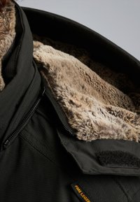 Giacca nera con un morbido rivestimento in finta pelliccia color beige. Caratterizzata da un colletto alto, chiusura con zip e dettagli in velcro sui bordi. Tessuto testurizzato visibile.