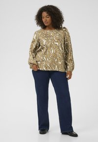 Blusa a maniche lunghe di colore marrone chiaro con un motivo ondulato bianco, abbinata a jeans dritti blu scuro e scarpe nere. Tessuto con una superficie liscia.
