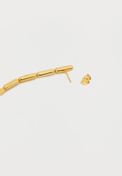 Χρυσή κυλινδρική αλυσίδα με χάντρες, με το ένα άκρο να έχει ευθύ καρφί και ξεχωριστό κλιπ πεταλούδα για σκουλαρίκι, σε λευκή επιφάνεια.