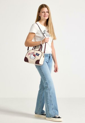 Junge Frau trägt ein weißes Hello Kitty T-Shirt und hellblaue Jeans, trägt eine beige Hello Kitty Handtasche mit dunkelroten Trägern.