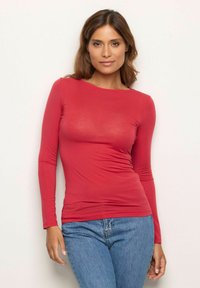 Mujer con cabello castaño claro vistiendo una camiseta ajustada de manga larga roja y jeans azules, de pie contra un fondo claro y sencillo.